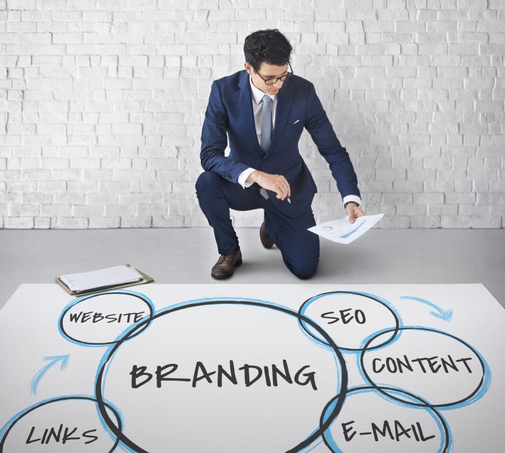 A man in a suit kneels on the floor, reviewing papers, near a diagram on branding strategies including website, SEO, content, links, and email, against a white brick wall.