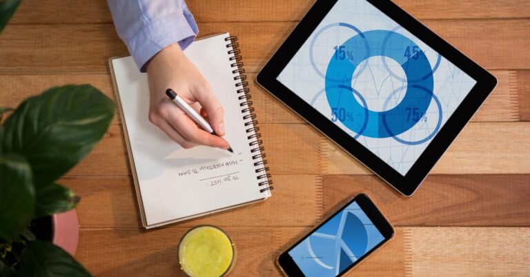 A person writes in a notebook on a wooden desk with a tablet displaying a pie chart, a smartphone, a plant, and a glass of juice nearby.