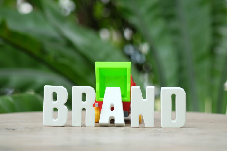 White 3D letters spelling the word ‘BRAND’ placed on a wooden surface with a small colorful toy truck in the background and green leaves blurred behind.